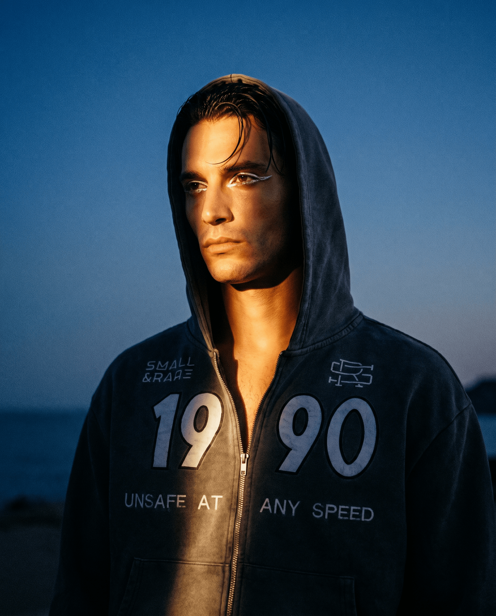 Product shot of a man wearing Grey 90 Zip Hoodie with the hoodie up featuring a bold "1990" and "Small And Rare" insignias on a beach setting.