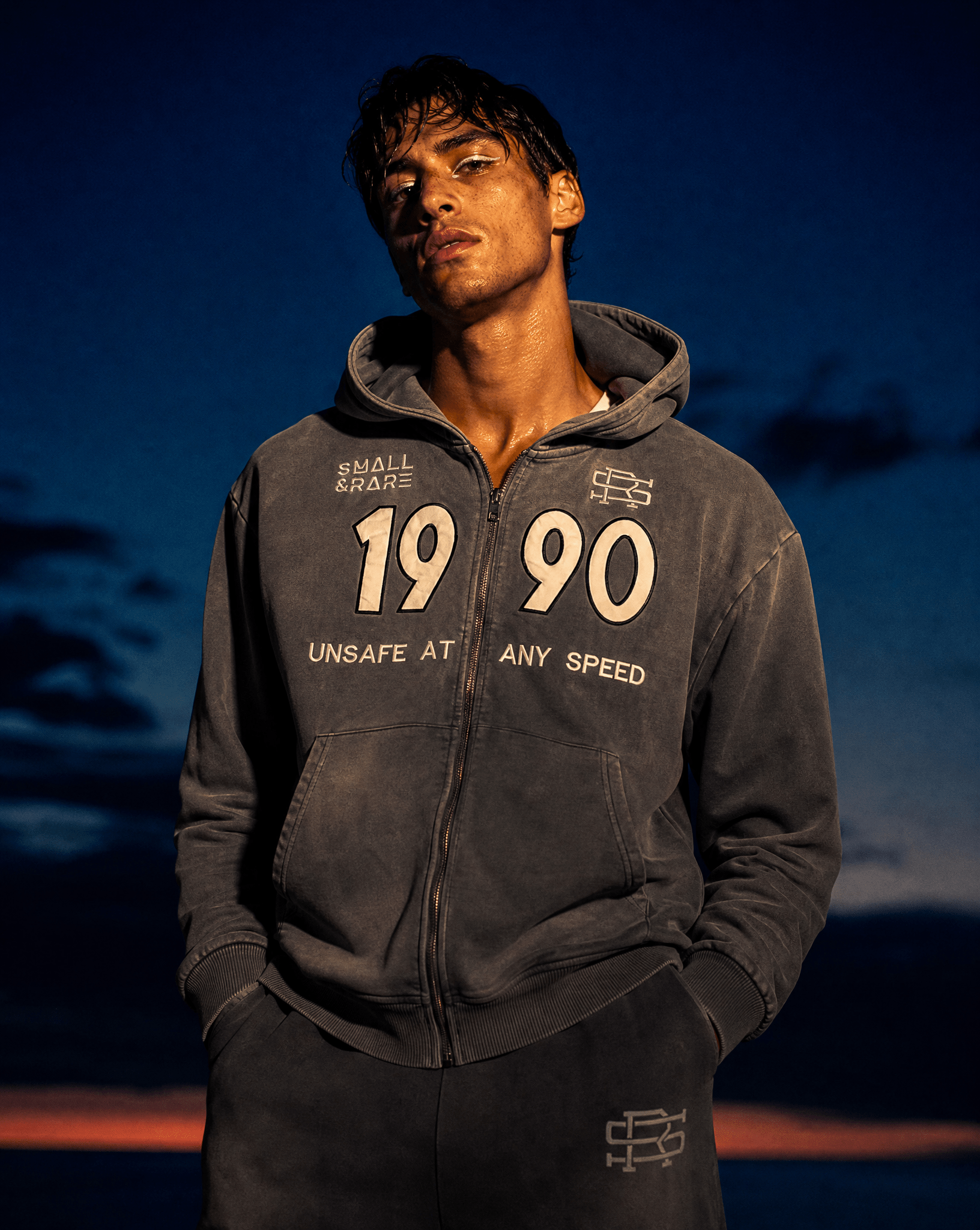 Product shot of a man wearing Grey 90 Sweatpants and Grey 90 Zip Hoodie featuring a bold "1990" and "Small And Rare" insignias on a beach setting.