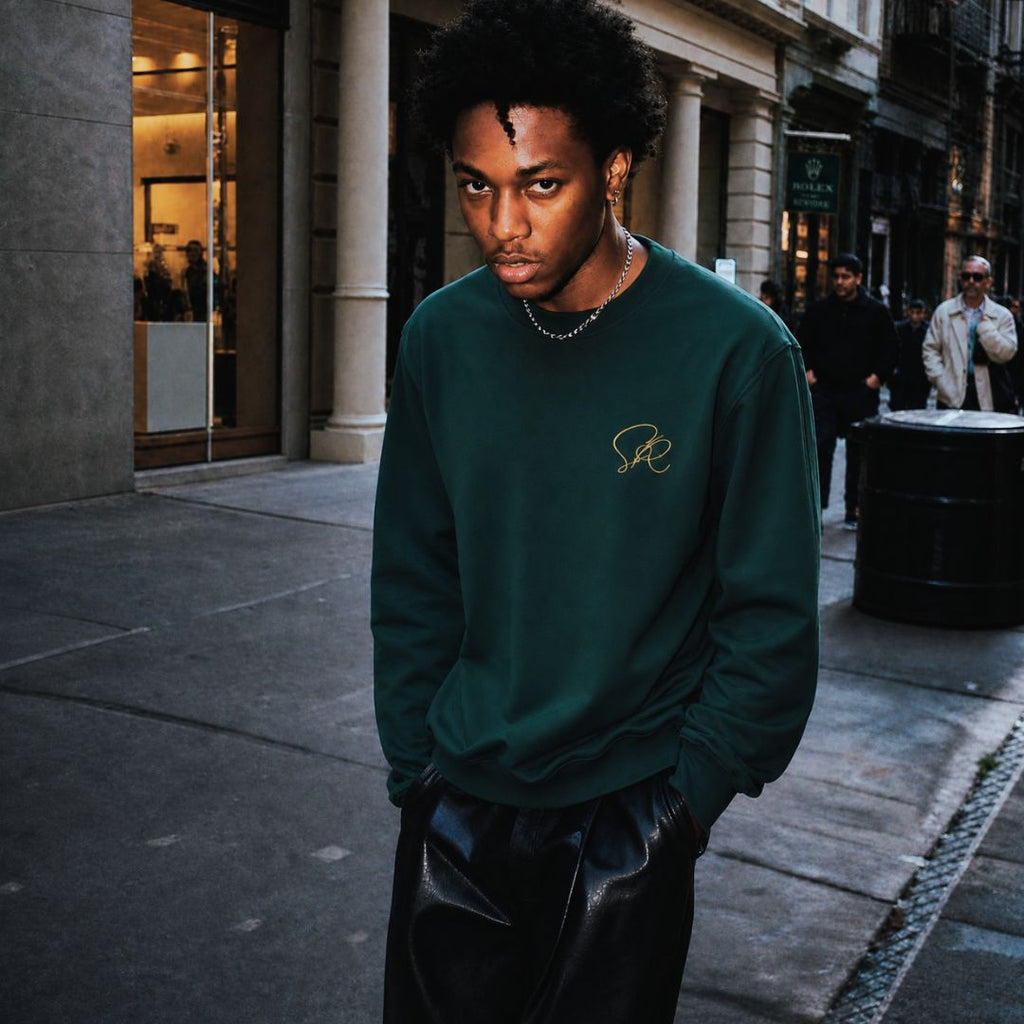 Product shot of man wearing Oversized Green Crewneck Sweatshirt featuring green embroidered insignia on left chest standing on the side of a sidewalk in a bustling city.