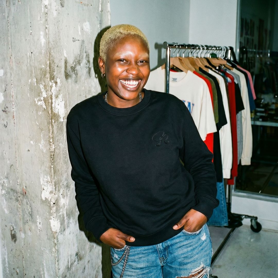 Product shot of woman wearing Black Core crewneck sweatshirt featuring black embroidered insignia on left chest standing in-front of a clothing rack leaning on a concrete wall.