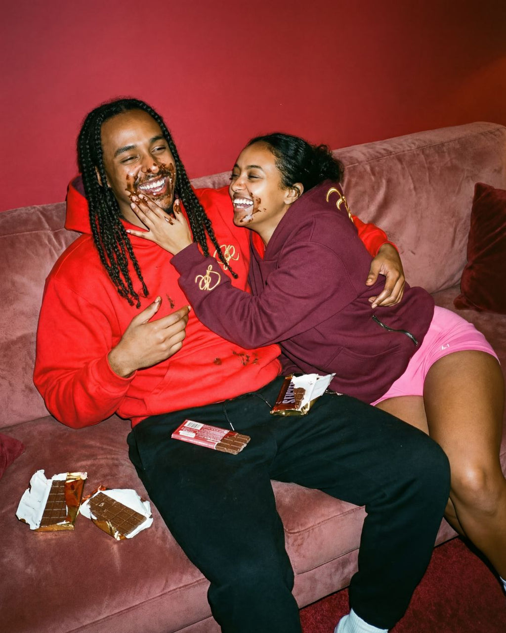 Two people sitting on a couch laughing, with one person wearing the red side of hoodie  and the other wearing the crimson side of the Crimson Blaze hoodie.