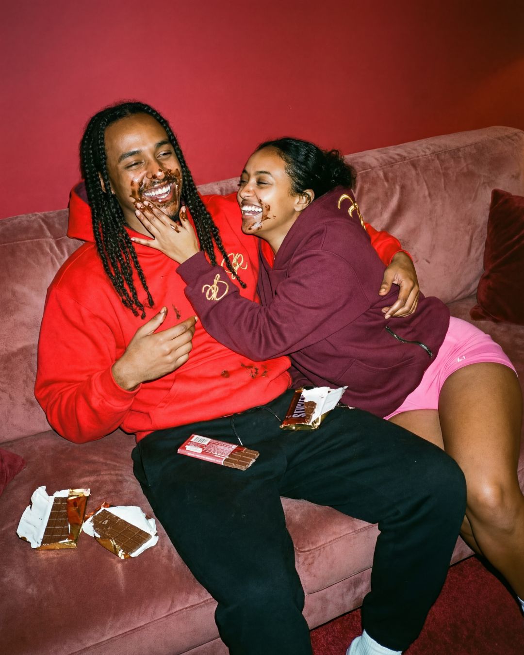 Two people sitting on a couch laughing, with one person wearing the red side of hoodie  and the other wearing the crimson side of the Crimson Blaze hoodie.